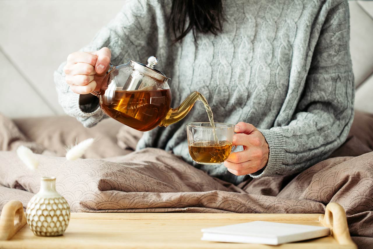 Woman dressed in knitted sweater pours hot tea from glass teapot into mug while sitting in bed